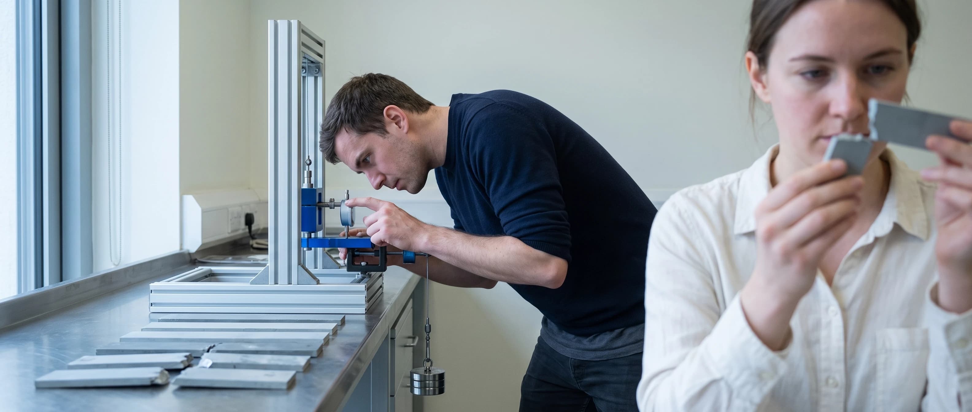 An engineer adjusting a test rig while a colleague inspects a machined sample