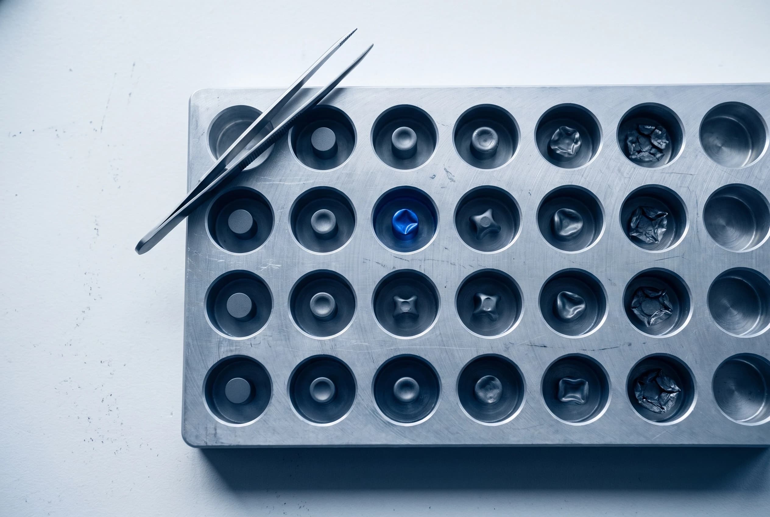 A metal tray of test wells with tweezers, one sample in blue