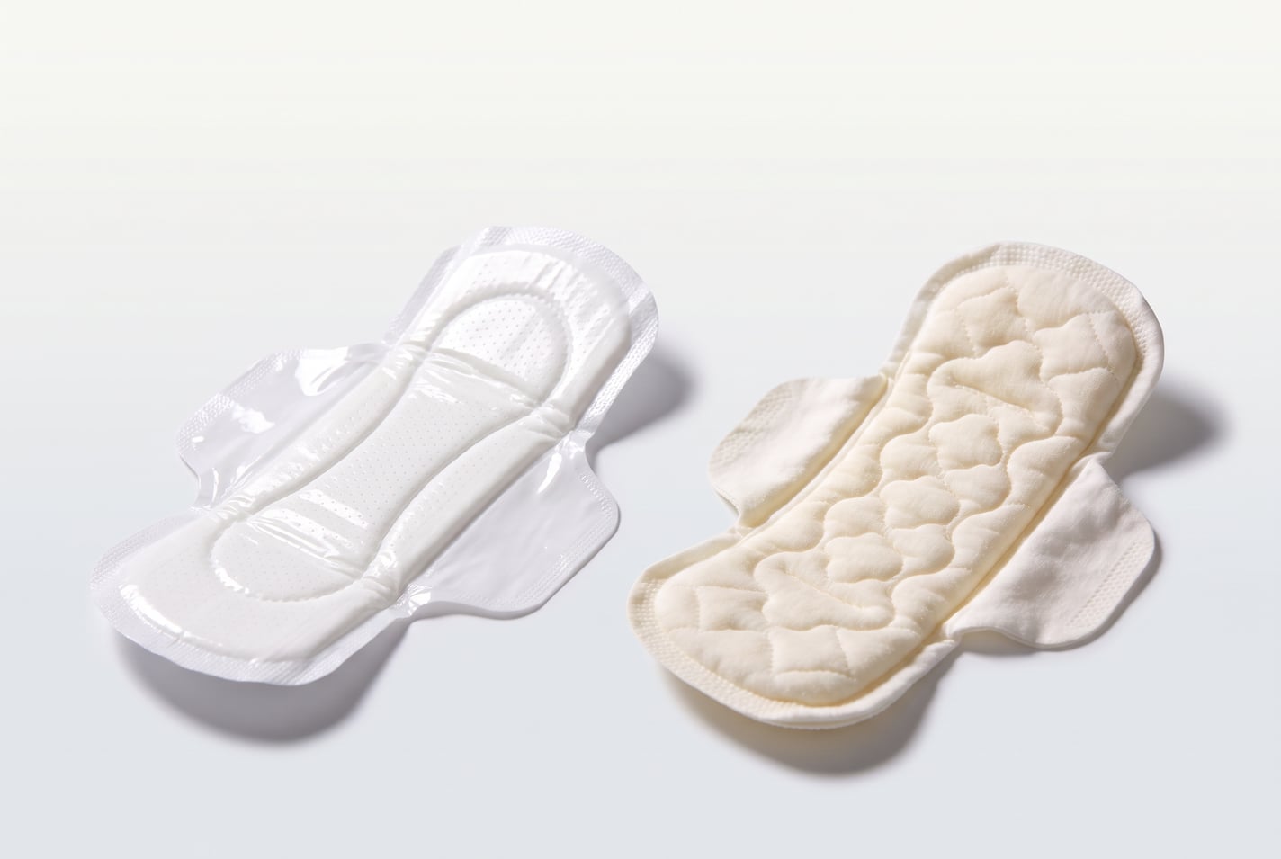 Render of two pads side by side on a pale ground: one with a glossy, perforated plastic-film topsheet, and the Alvera pad with its matte quilted topsheet