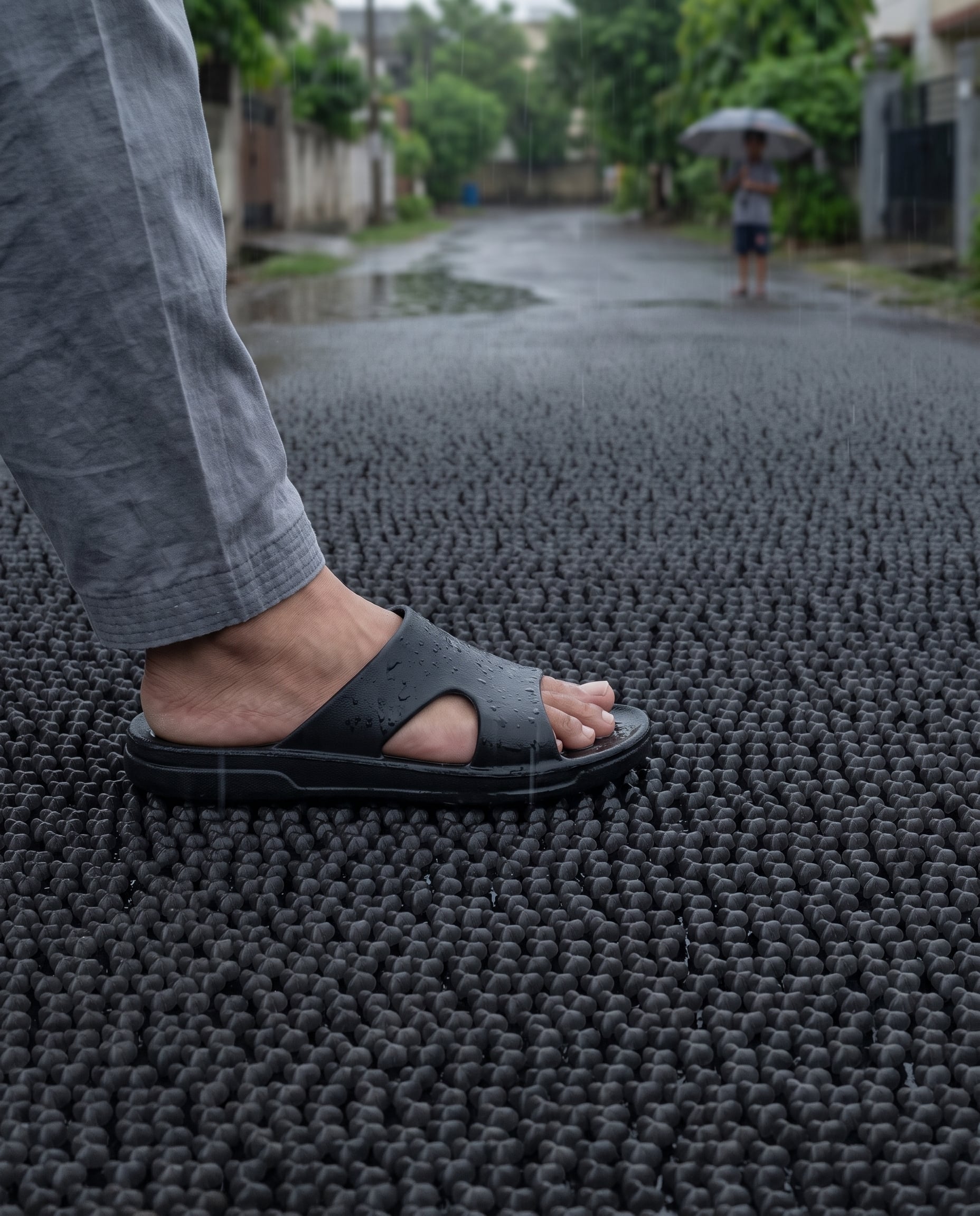 Render of a foot in a black sandal stepping onto a rain-wet street surfaced with black Oberon modules, a figure under an umbrella further down the lane